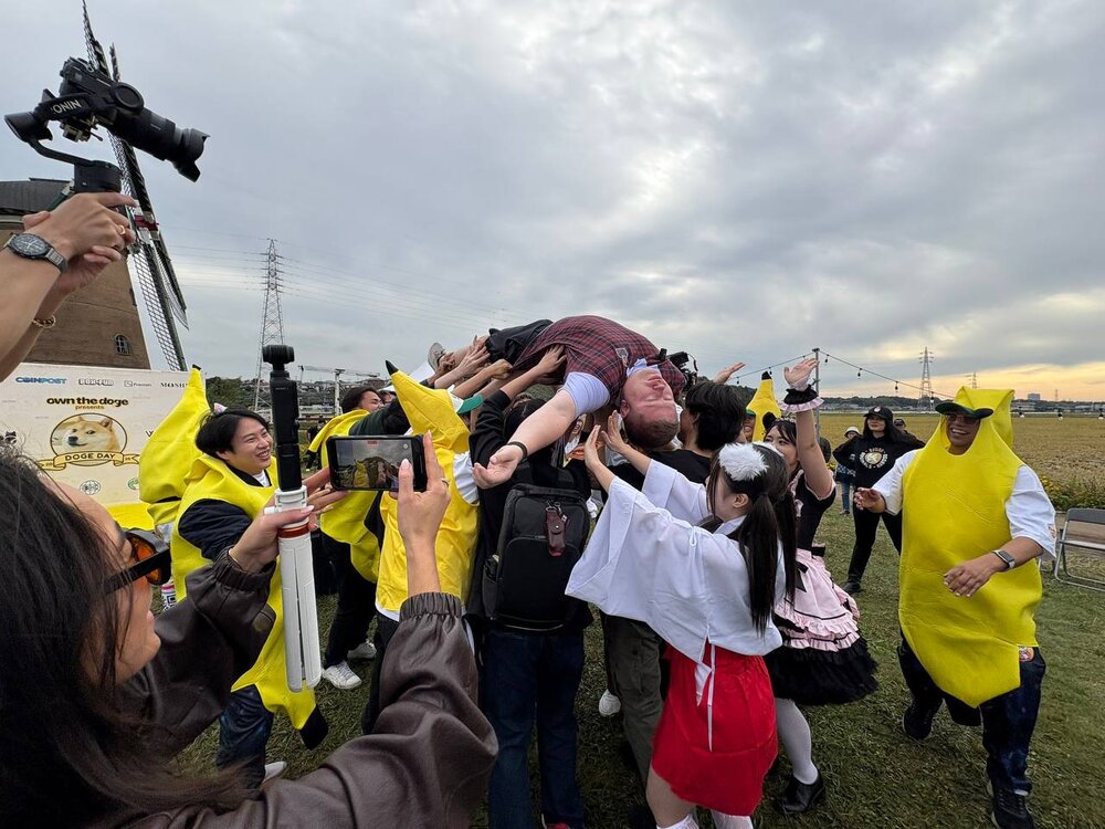 A man held up on his back by a crowd of fans in banana, shrine maiden and maid costumes, with people filming on phones and a gimbal camera in front of the windmill