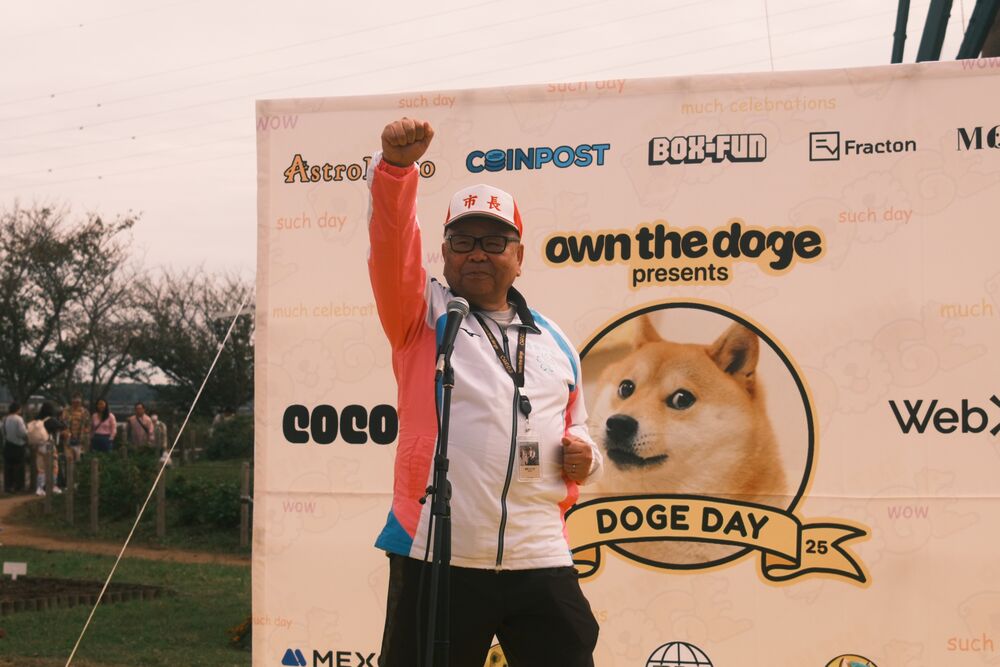 An organizer raising a fist at the microphone in front of the Own The Doge Doge Day backdrop