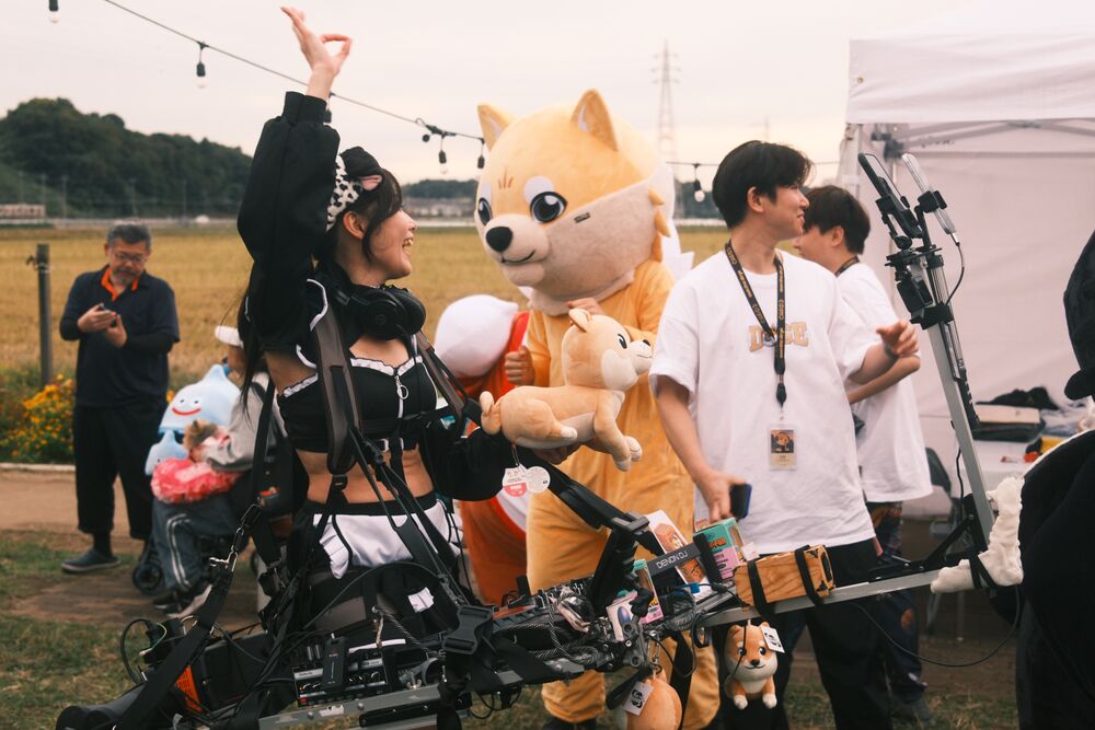 A Shiba mascot holding a plush dog beside a performer and a camera crew