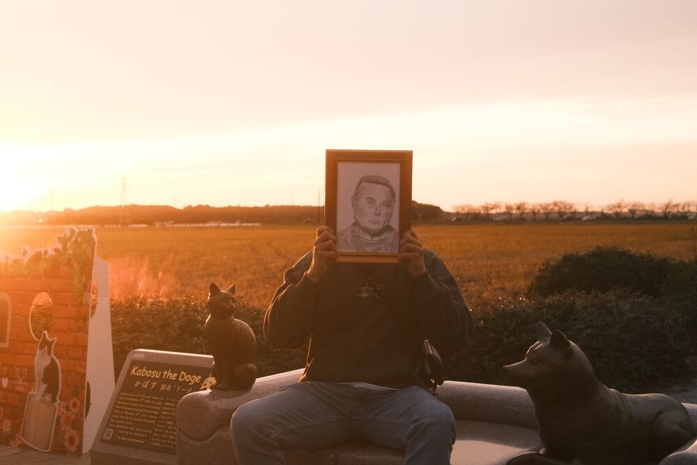 A visitor seated at the Kabosu statue at golden hour, holding a framed portrait in front of their face