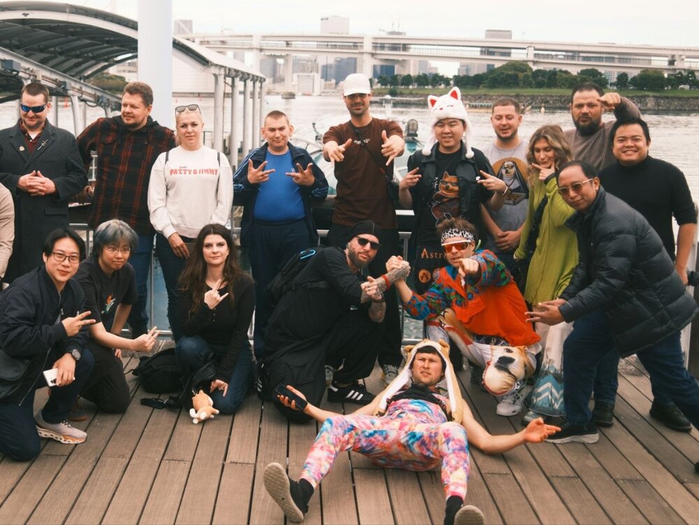 About eighteen people crowded onto a wooden pier in front of a harbor and a highway bridge, flashing hand signs and pointing at the camera, with one in a colorful tie-dye suit and a Shiba hood sprawled across the front