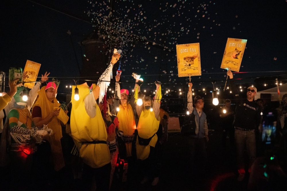 Fans in banana costumes cheering under string lights at night as bubbles drift overhead, holding up Doge Day signs