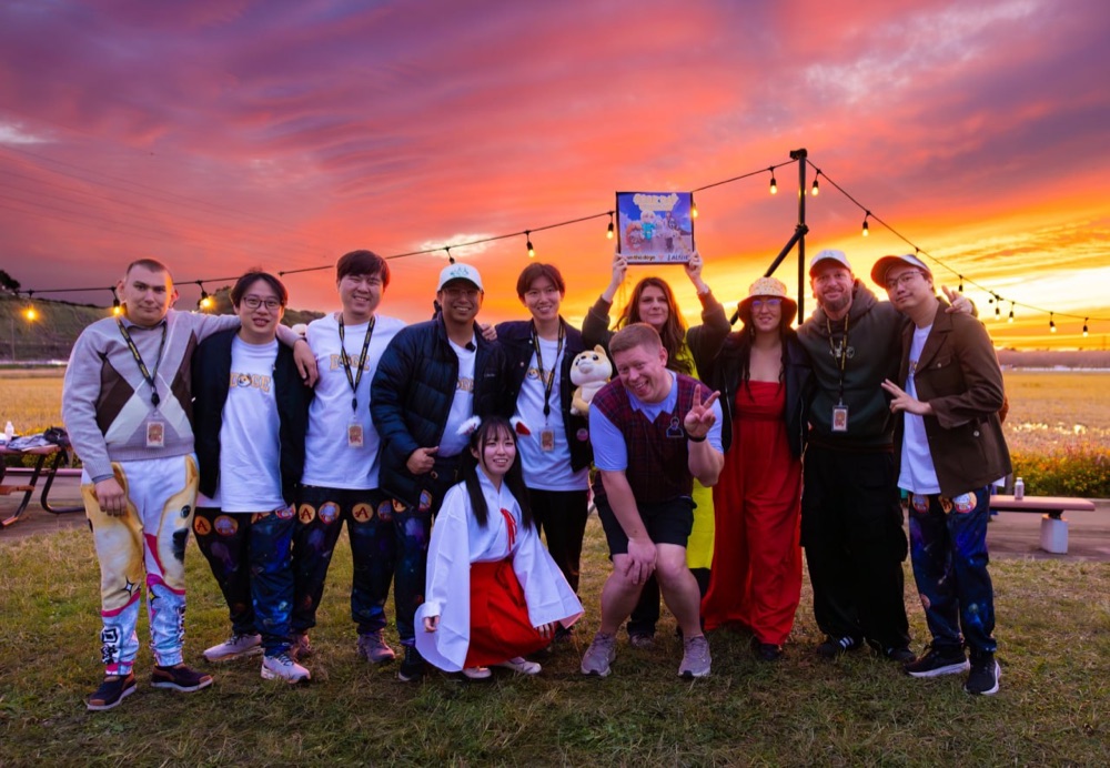 The organizing team arm in arm on the grass under a blazing orange and purple sunset, one holding up a framed Doge Day artwork